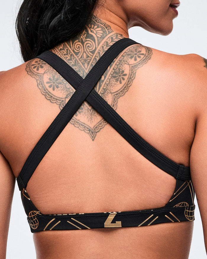 Close-up back view of a female model wearing a black cross-strap bra with a geometric pattern and gold accents on a plain background.