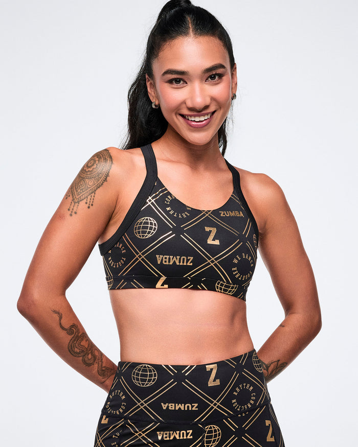 Female model wearing a black Zumba sports bra with gold logos and matching leggings on a white background.