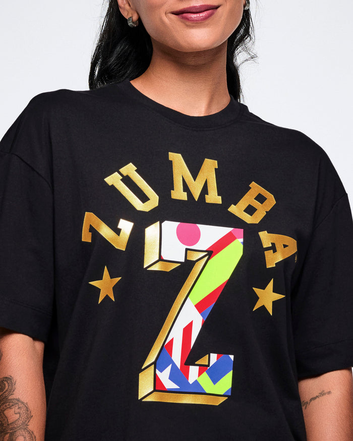 Close-up of a female model wearing a black Zumba T-shirt featuring a colorful flag-patterned big “Z” and arched gold “Zumba” text with gold stars above the big Z.