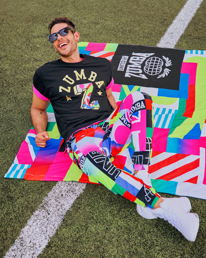 Male model in sunglasses chilling on a grassy field wearing a black t-shirt, colorful Zumba pants and white shoes.