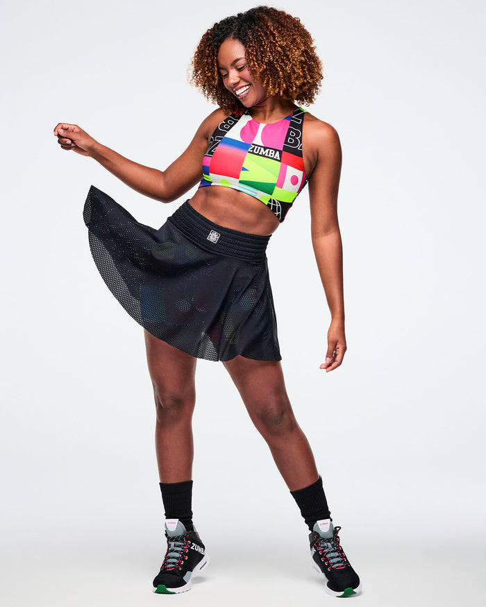 Female model wearing a colorful sports bra, black mesh skort, black socks and black shoes on a white background.