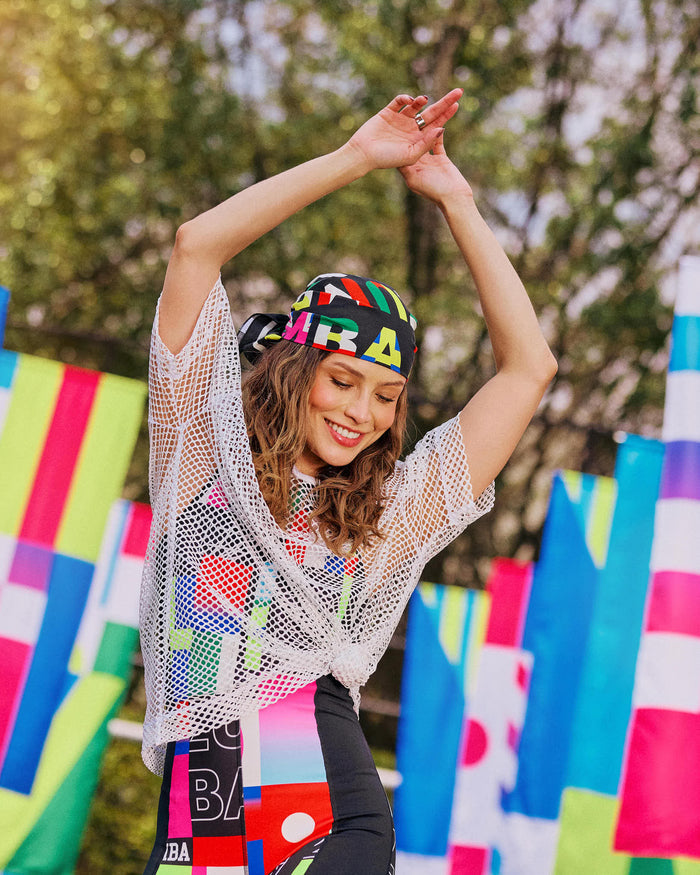 Female model dancing outdoors with arms raised, a bandana on her head, wearing a silver mesh top over a multicolored bodysuit.