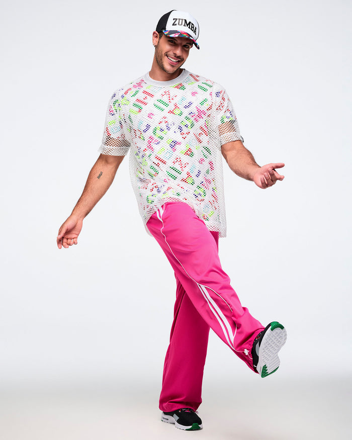 Male model posing with a forward kick, wearing a silver mesh top over a colorful Zumba shirt, pink pants, black shoes, and a Zumba hat.