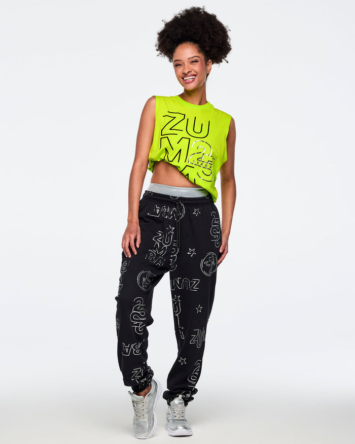 Neon green sleeveless Zumba tank with oversized black logo graphics and black jogger pants covered in silver Zumba prints. Female model wears the bright muscle tank with loose black drawstring joggers and silver athletic sneakers, smiling in a full-body pose against a light gray background.