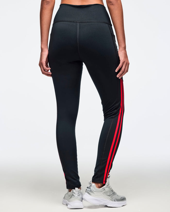 Black high-waisted leggings shown from the back with a sleek fitted silhouette, side pockets, and double coral pink side stripes. Female model is wearing the full-length leggings with silver athletic sneakers.