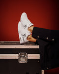 A pair of white low-top sneakers with black graphic details and the brand name 'ZUMBA' visible on the side, worn with black pants and posed on top of a black equipment case against a dark red background.