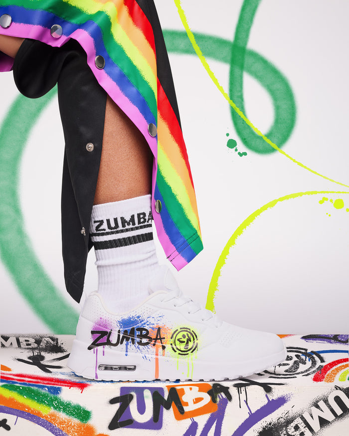 Side view of a white sneaker with colorful graffiti-style accents and “Zumba” in black text, worn with white Zumba socks and black pants featuring a rainbow stripe, set against an abstract graffiti-style background.
