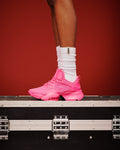 A bright pink shoe worn with white crew socks, shown on a female model's foot standing on top of a black equipment case against a solid red background.
