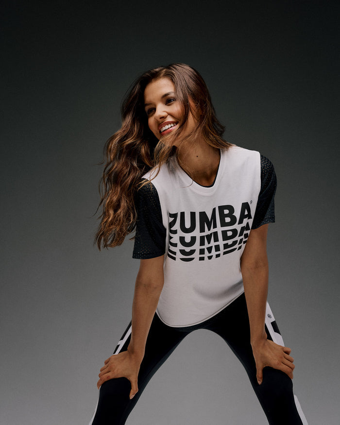 Female model wearing a black and white 'Zumba' outfit with hands on her knees smiling.