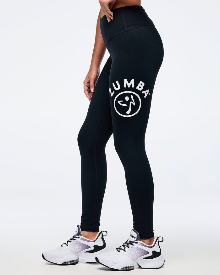 A female model wearing black high-waisted Zumba leggings with a white Zumbito logo on the left thigh, paired with white and black athletic sneakers, shown from a side profile.