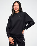 Female model wearing the black waffle-knit crewneck sweatshirt, with a white ZUMBA text on the upper left and paired with matching pants, one hand resting on her hip.