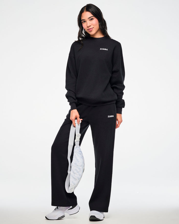 Black waffle-knit crewneck sweatshirt and matching sweatpants with a small white 'ZUMBA' logo, worn by a female model carrying a white 'ZUMBA'-print crossbody bag.