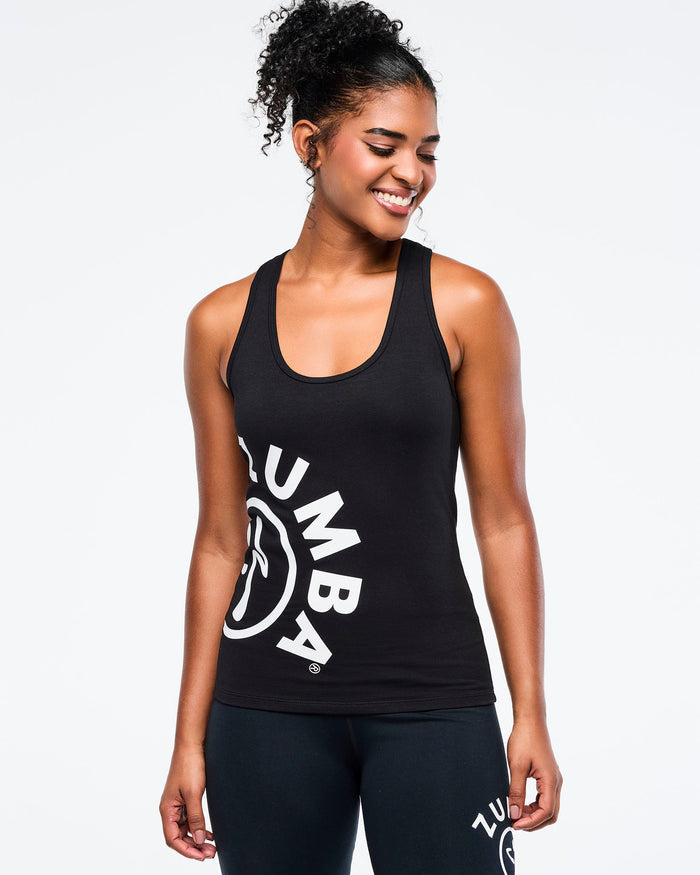 Female model wearing a black Zumba tank with arched “ZUMBA” text and a large white Zumbito graphic on the right side paired with matching biker shorts.