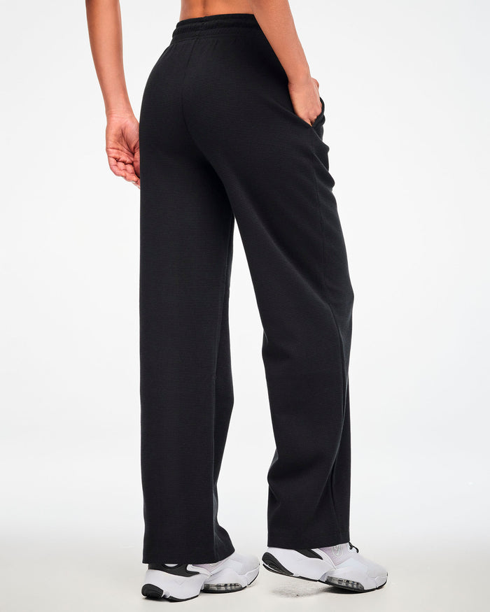 Back view of the black waffle-knit wide-leg track pants, female model with one hand in a side pocket.