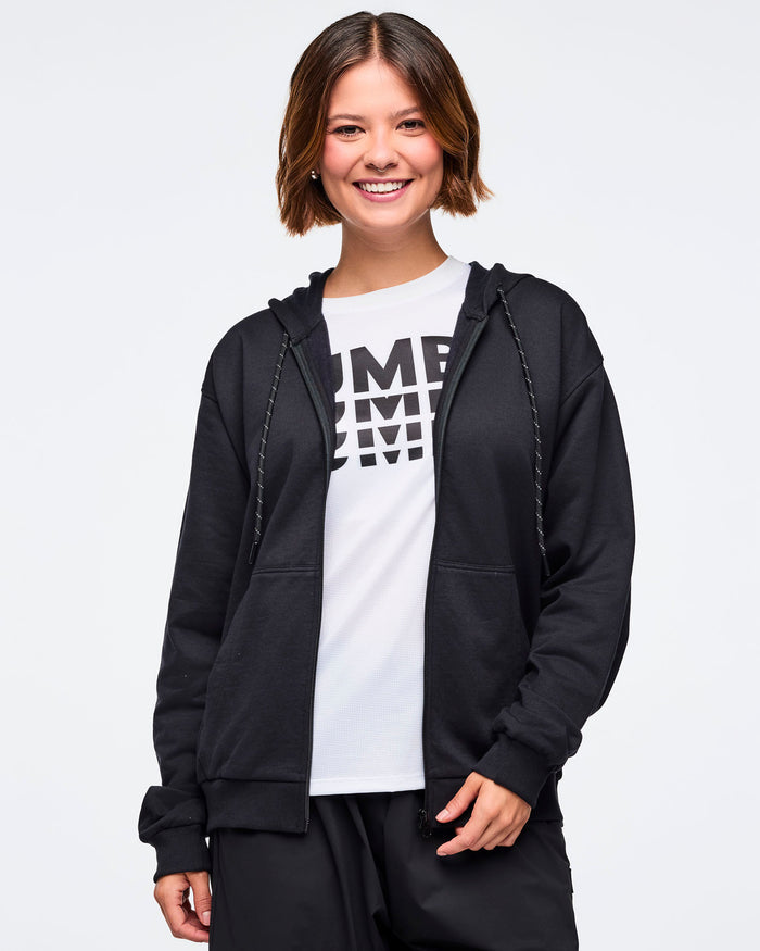 Front view of a female model wearing an open black zip-up hoodie, layered over a white "ZUMBA" graphic tee, paired with black pants, against a white background.