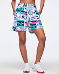 White elastic-waist drawstring shorts with an all-over teal-and-pink Zumba beach graphic print featuring waves, sunglasses, anchors, flowers, and logos; shown on a female model with white sneakers with hot-pink soles.