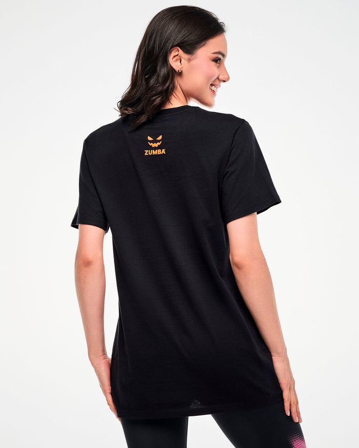 Shown from the back, female model wearing a black tee with a small orange jack-o'-lantern graphic and "ZUMBA" text.