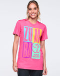 Bright pink Zumba T-shirt with oversized multicolor “ZUMBA” graphic and “BOSS MOVES” text on the front, worn by a female model with a crew neckline and short sleeves, paired with black pants.