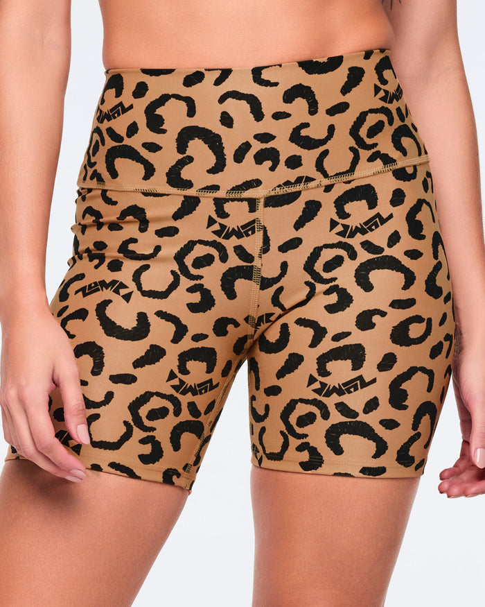 Close-up of tan leopard-print biker shorts with black abstract spots and logo accents, featuring a wide high-rise waistband and smooth fitted silhouette, shown on a female model.