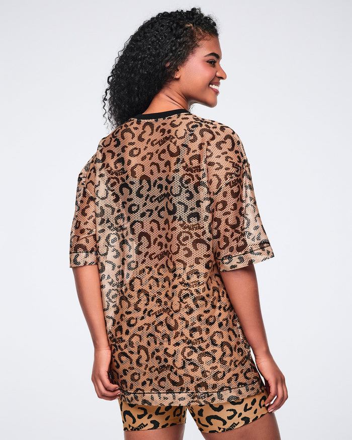 Tan leopard-print mesh top with black abstract spots, a black V-neck collar, oversized fit, and elbow-length sleeves, shown from the back on a smiling female model wearing matching leopard-print biker shorts.
