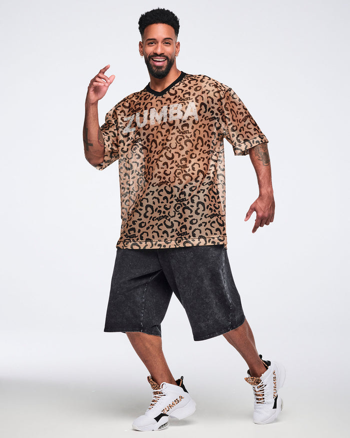 Tan leopard-print mesh top with black abstract spots, a black V-neck collar, oversized fit, and elbow-length sleeves, shown on a smiling male model wearing black acid-wash long shorts and white high-top sneakers with leopard-print details.