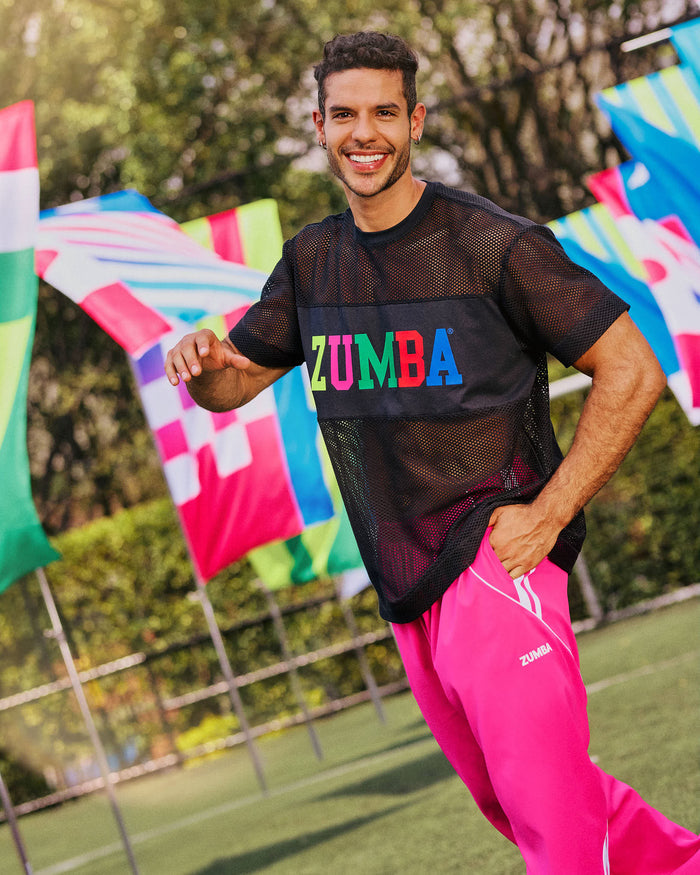 Male model wearing a Zumba black mesh shirt and pink pants on a grassy field with colorful flags in the background.