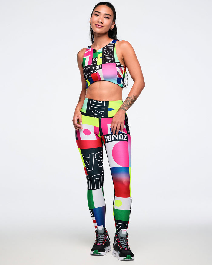 Female model wearing a colorful sports bra and matching leggings, paired with black Zumba shoes against a white background.